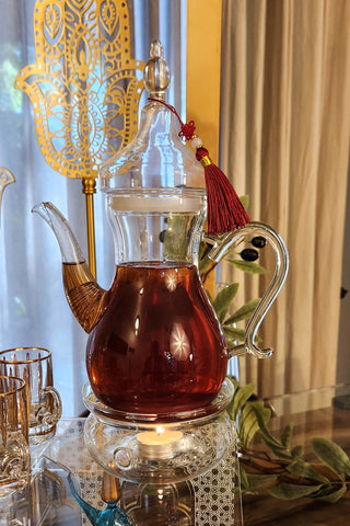 Image Ibreeq a turkish style teapot with tea inside next to Anater food riser and tea cups and green leaves background.